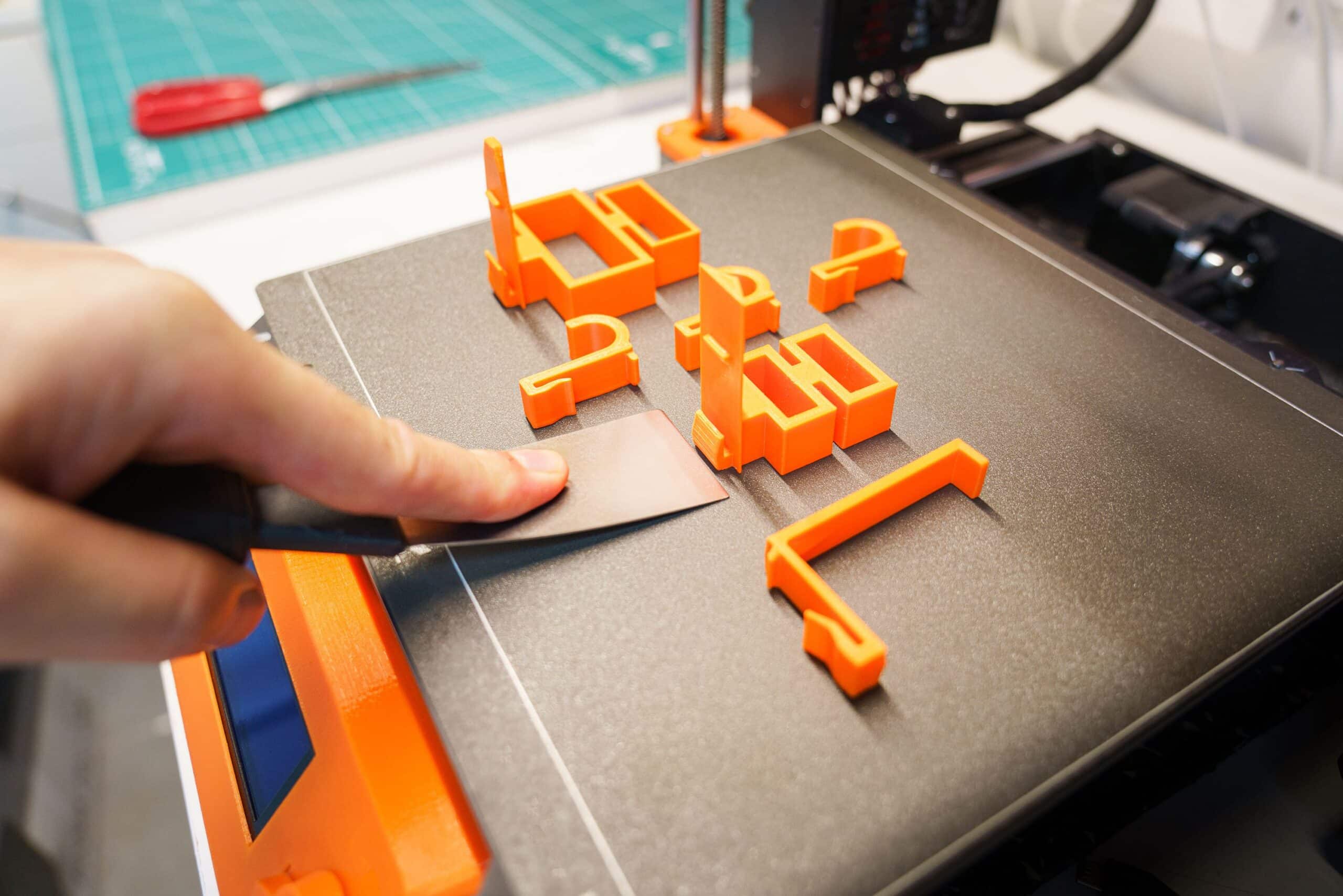 A hand carefully removes an orange 3D printed object from a printer bed using a spatula